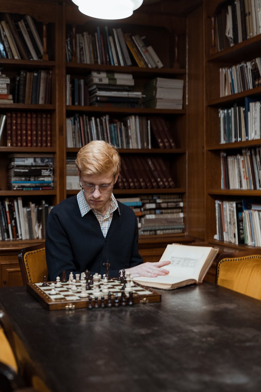 man sitting at table with chess while holding a book